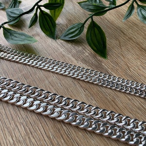 Könnte beinhalten: Zwei silberne Curb-Ketten in verschiedenen Längen auf einer Holzoberfläche. Die Ketten bestehen aus dicken, ineinandergreifenden Gliedern.