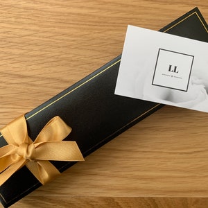 Puede incluir: Una caja de regalo negra con una cinta dorada y una tarjeta blanca con el texto "LL" en ella.