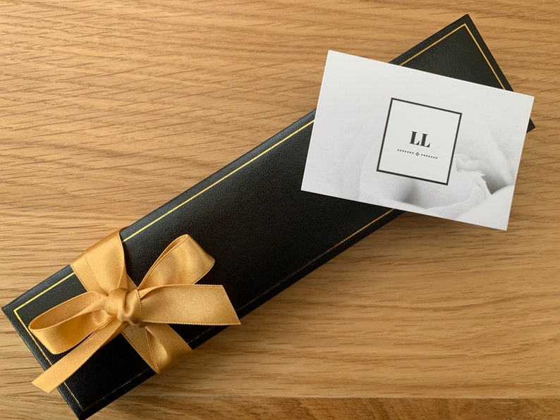 Puede incluir: Una caja de regalo negra con una cinta dorada y una tarjeta blanca con el texto "LL" en ella.