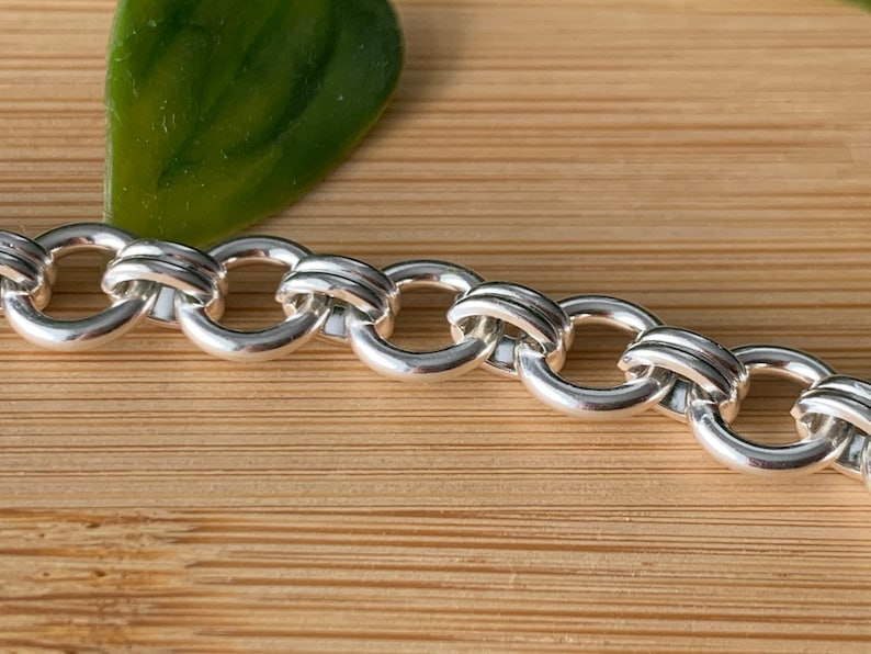 Puede incluir: Un brazalete de cadena de plata con un dise&ntilde;o &uacute;nico e entrelazado. La cadena est&aacute; hecha de eslabones redondos y gruesos que est&aacute;n conectados de una manera que crea una serie de peque&ntilde;os c&iacute;rculos superpuestos.