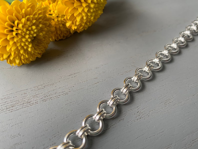 Puede incluir: Un brazalete de cadena de plata con un dise&ntilde;o entrelazado &uacute;nico. La cadena est&aacute; hecha de varios c&iacute;rculos peque&ntilde;os que est&aacute;n unidos para crear un brazalete resistente y duradero.