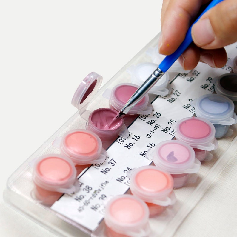 May include: A paint-by-numbers kit with small, numbered paint pots in shades of pink and purple. A hand holds a paintbrush with a blue handle, dipping it into a pot of pink paint. The kit includes a numbered guide.