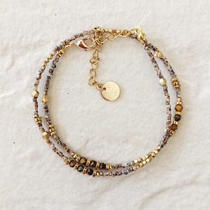 Peut inclure: Un bracelet à plusieurs rangs avec deux rangs de petites perles multicolores et de perles métalliques dorées. Le bracelet a un fermoir doré et un petit charme rond. Les perles sont marron, beige et dorées.