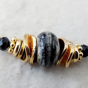 Puede incluir: Un collar de cuentas con una cuenta central jaspeada en gris y negro, flanqueada por espaciadores y discos de metal dorado. El diseño incluye pequeñas cuentas negras facetadas y cuentas metálicas doradas dispuestas en un patrón simétrico.