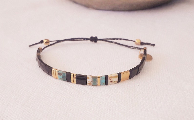 May include: A black and gold beaded bracelet with a black cord and gold beads. The bracelet has a small gold charm attached to it.