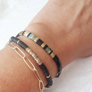 May include: Three bracelets stacked on a wrist. The top bracelet is black with gold and turquoise accents. The middle bracelet is black with gold accents. The bottom bracelet is a gold chain.