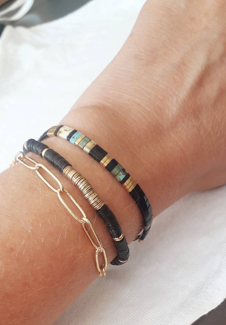 May include: Three bracelets stacked on a wrist. The top bracelet is black with gold and turquoise accents. The middle bracelet is black with gold accents. The bottom bracelet is a gold chain.