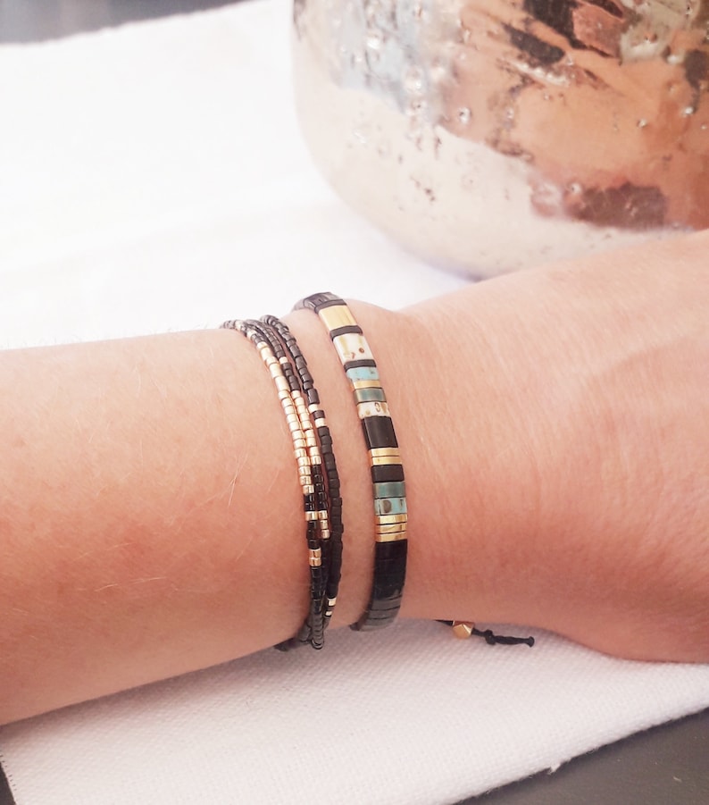 May include: A set of three bracelets with black, gold, and turquoise beads. The bracelets are stacked on a person's wrist.