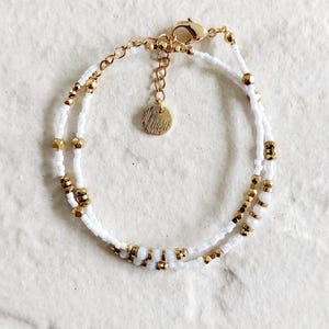 Op de afbeelding: Een delicate armband met witte zaadkralen en goudkleurige metalen kralen. De armband heeft een dubbelstrengs ontwerp met een goudkleurige sluiting en een kleine, ronde bedel. De armband is ongeveer 17.8 cm lang.