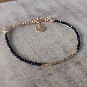 Puede incluir: Un brazalete dorado con cuentas negras y cuentas cuadradas doradas. El brazalete tiene un cierre dorado y un pequeño disco de oro.