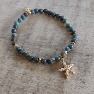 May include: A beaded bracelet featuring blue and brown spherical beads with gold-colored accents. A gold starfish charm hangs from the bracelet. The bracelet is displayed on a wooden surface.