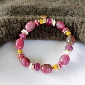 May include: A beaded bracelet featuring a mix of pink, white, yellow, and gold-colored beads. The bracelet has large, oval-shaped pink beads interspersed with smaller, rounder beads in various colors. The clasp is gold-colored.