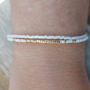May include: Two bracelets on a wrist. The top bracelet is white and the bottom bracelet is gold.