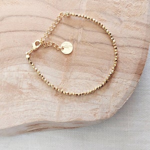 May include: A delicate gold bracelet with small, faceted beads. The bracelet has an adjustable chain and a small, round charm. The jewellery is displayed on a light-coloured wooden surface.