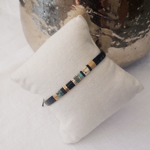 May include: A black and gold beaded bracelet with turquoise and white beads. The bracelet is on a white fabric surface.