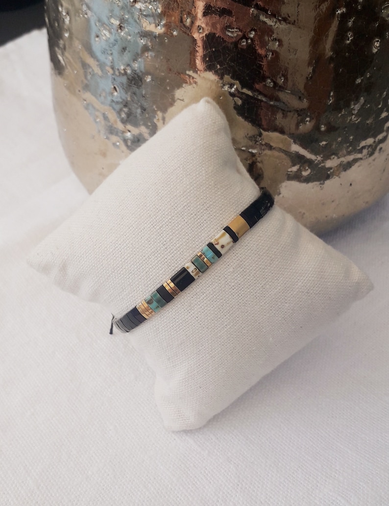 May include: A black and gold beaded bracelet with turquoise and white beads. The bracelet is on a white fabric surface.