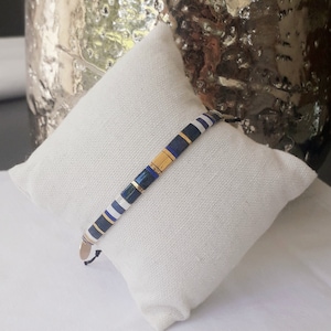 May include: A blue, white and gold beaded bracelet with a gold clasp. The bracelet is on a white fabric surface.