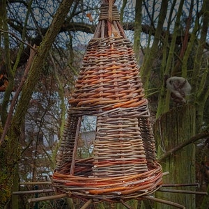 Könnte beinhalten: Handgefertigter Vogelhaus-Futterspender in konischer Form, gewebt aus braunen und rotbraunen Weidenzweigen. Der Futterspender wird an einem Seil aufgehängt und hat eine kreisförmige Sitzstange aus kleinen Zweigen.