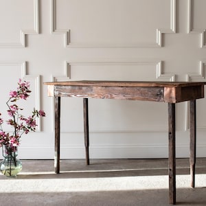 May include: A rustic wooden table with a distressed finish, featuring four slender legs. The tabletop is a warm brown, and the legs are a darker shade. A vase of pink flowers sits to the left, adding a touch of color. The table is approximately 36 inches long.