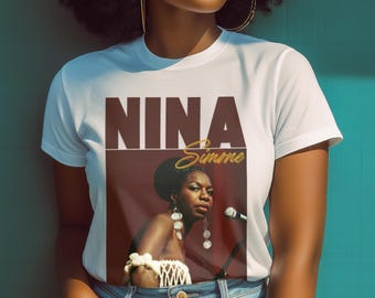 May include: White t-shirt featuring a portrait of Nina Simone with the name "NINA" in a dark brown font and "Simone" in gold script. The image on the shirt shows Simone singing into a microphone, wearing earrings and a white top.