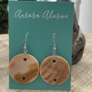 May include: A pair of round, beige earrings with a marbled pattern, suspended from silver-colored hooks. The earrings are displayed on a teal card with the text "Aurora Alarice". The background is a neutral beige fabric.