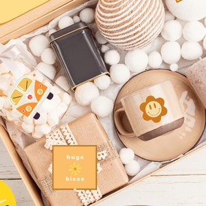 Puede incluir: Una caja de regalo llena de varios artículos, incluyendo una taza con una cara sonriente, una lata pequeña, un frasco de malvaviscos y una etiqueta que dice "hugs kisse". La caja está decorada con un sol y las palabras "Sunshine" y "bad days don't stay bad forever".
