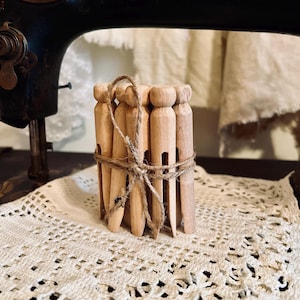 May include: A bundle of unfinished wooden clothespins tied together with twine. The clothespins are light brown and cylindrical with a slot for holding clothes. The background includes a vintage Singer sewing machine and a white lace doily.