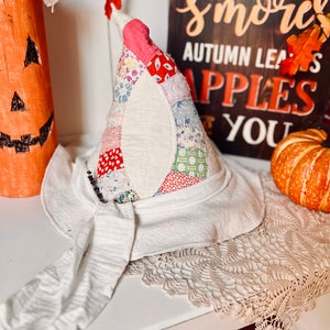 May include: A handmade witch hat with a patchwork cone in red, pink, and floral patterns, atop a white brim with a long, draped tie. The hat is part of a fall-themed display, with a pumpkin and a sign that reads "Bonfires, S'mores, Autumn Leaves, Apples, You."