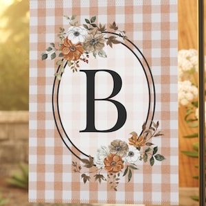 Könnte beinhalten: Gartenflagge mit einem Vichy-Muster in Pfirsich- und Weißtönen. Die Flagge zeigt einen großen schwarzen Buchstaben "B" in einem ovalen Rahmen, umgeben von floralen Arrangements in gedämpften Farben. Auf einem schwarzen Metallständer.