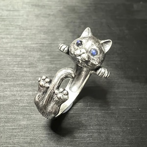 May include: A silver ring featuring a detailed cat design. The cat has blue eyes and is holding onto the ring band with its paws.