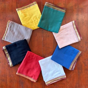 Może przedstawiać: Osiem kwadratów tkaniny w różnych kolorach, w tym żółtym, zielonym, szarym, czarnym, czerwonym, niebieskim, białym i różowym, ułożonych w okrąg na drewnianej powierzchni. Każdy kwadrat ma złotą i czerwoną lamówkę.