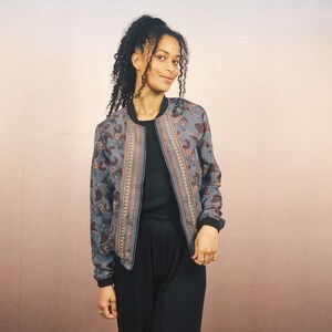 May include: A blue bomber jacket with a paisley pattern in brown and orange. The jacket has a black collar, cuffs, and hem. The jacket is open, revealing a black top. The background is a gradient of pink and beige.