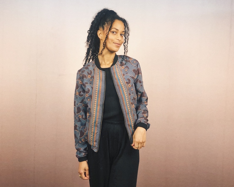 May include: A blue bomber jacket with a paisley pattern in brown and orange. The jacket has a black collar, cuffs, and hem. The jacket is open, revealing a black top. The background is a gradient of pink and beige.