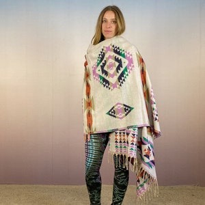 May include: A white woven shawl with a geometric pattern in shades of pink, orange, green, and blue. The shawl has fringe on the edges and is draped over a person's shoulders.