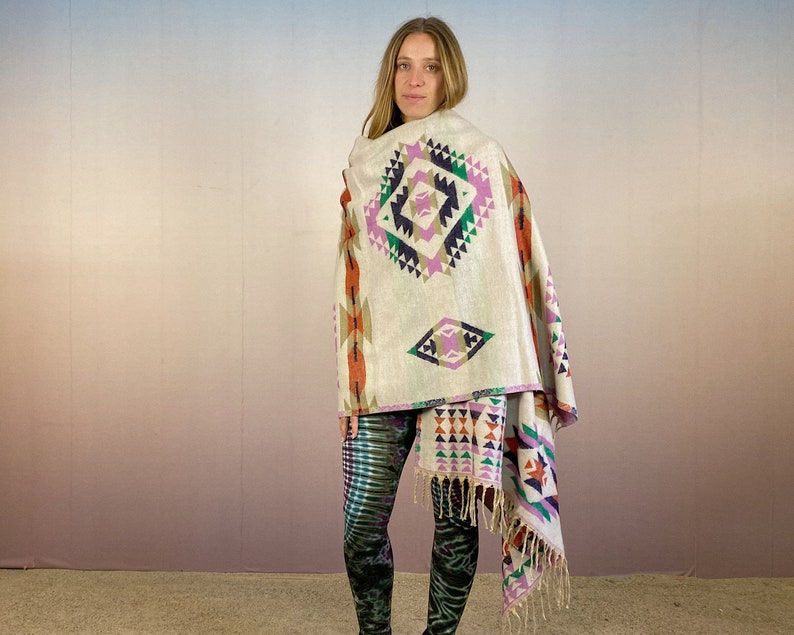 May include: A white woven shawl with a geometric pattern in shades of pink, orange, green, and blue. The shawl has fringe on the edges and is draped over a person's shoulders.