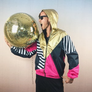 May include: A gold and black disco ball held by a person wearing a gold, black, pink, and white retro jacket with a hood. The jacket has a zipper and a striped design on the sleeves. The disco ball is round and reflective.