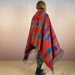 May include: A red, blue, and turquoise patterned throw blanket with fringe. The blanket is draped over a person's shoulders.