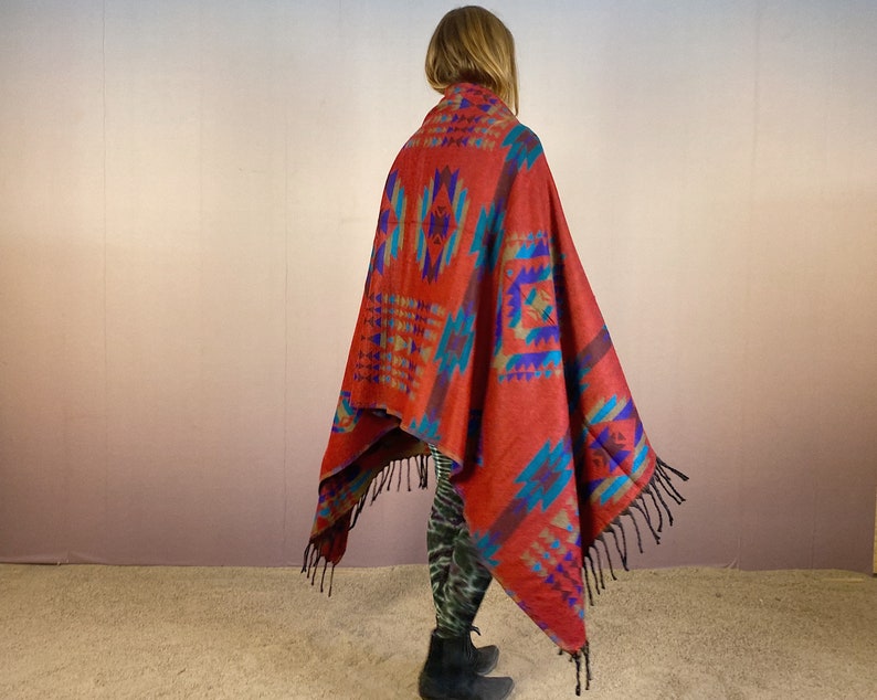May include: A red, blue, and turquoise patterned throw blanket with fringe. The blanket is draped over a person's shoulders.
