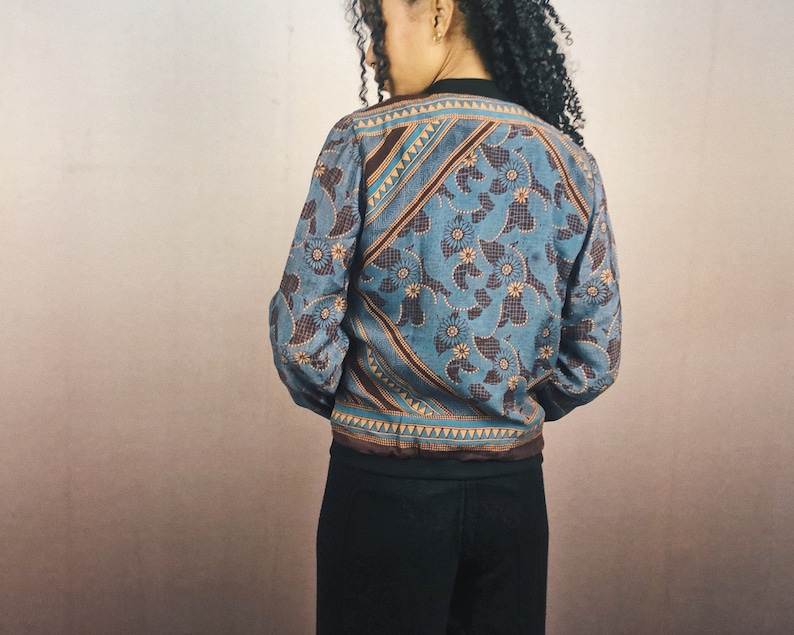 May include: A blue bomber jacket with a floral pattern and brown accents. The jacket has a black collar and cuffs. The floral design is in a lighter blue color, with brown and yellow details. The jacket is worn over black pants.