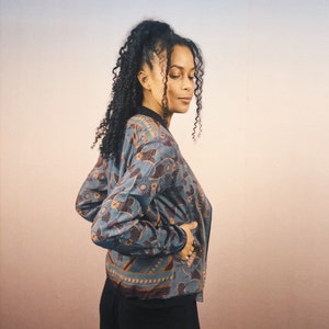May include: A blue and brown patterned bomber jacket with a black collar and cuffs. The jacket features a floral and geometric design. The jacket is worn by a person.