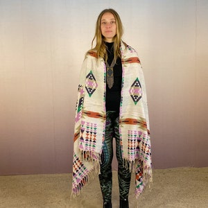 May include: A white woven shawl with a geometric pattern in shades of pink, orange, green, and blue. The shawl has fringe on the edges and is draped over a person's shoulders.