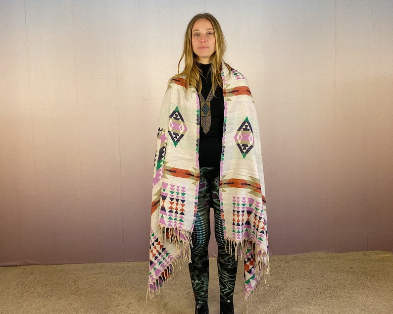 May include: A white woven shawl with a geometric pattern in shades of pink, orange, green, and blue. The shawl has fringe on the edges and is draped over a person's shoulders.