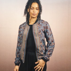 May include: A blue bomber jacket with a floral and geometric pattern in brown and orange. The jacket has a black collar and cuffs, and a full-length zipper. The jacket is worn over a black top.