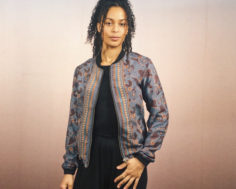 May include: A blue bomber jacket with a floral and geometric pattern in brown and orange. The jacket has a black collar and cuffs, and a full-length zipper. The jacket is worn over a black top.