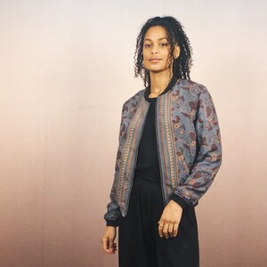 May include: A patterned bomber jacket in shades of blue, brown, and orange. The jacket features a floral design and a black collar and cuffs. The person is wearing a black top and black pants.