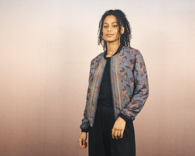 May include: A patterned bomber jacket in shades of blue, brown, and orange. The jacket features a floral design and a black collar and cuffs. The person is wearing a black top and black pants.