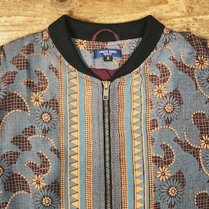 May include: A zip-up bomber jacket with a floral and geometric pattern in shades of blue, brown, and orange. The jacket has a black collar and a label that reads "TANTE EDEN BERLIN". The jacket is size small.
