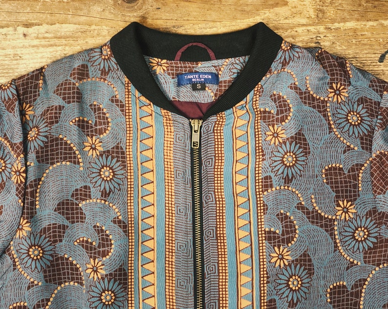 May include: A zip-up bomber jacket with a floral and geometric pattern in shades of blue, brown, and orange. The jacket has a black collar and a label that reads "TANTE EDEN BERLIN". The jacket is size small.