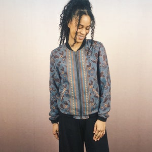 May include: A blue bomber jacket with a floral and paisley pattern. The jacket has a black zipper, cuffs, and collar. The pattern includes orange and brown accents. The jacket is worn with black pants.