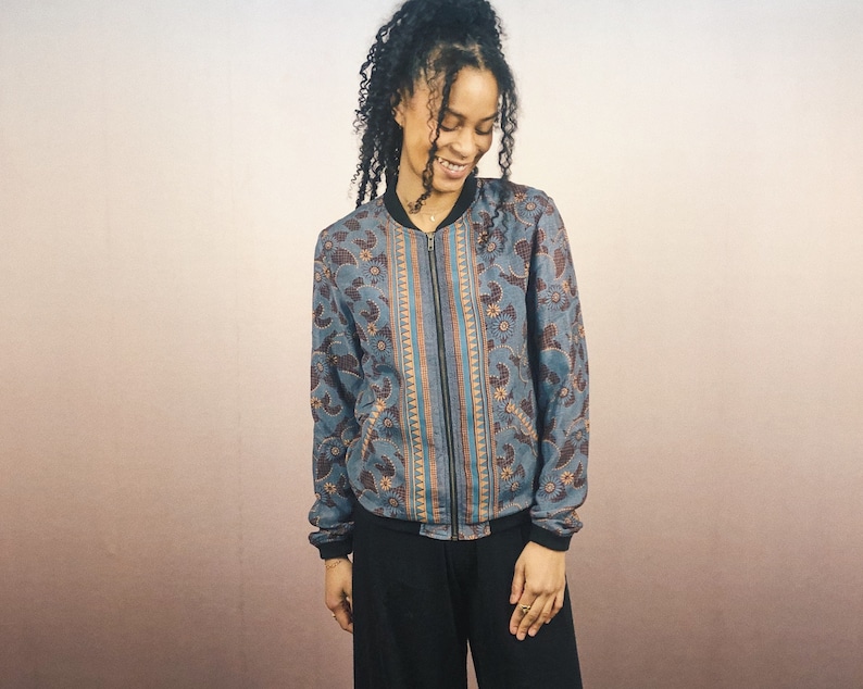 May include: A blue bomber jacket with a floral and paisley pattern. The jacket has a black zipper, cuffs, and collar. The pattern includes orange and brown accents. The jacket is worn with black pants.